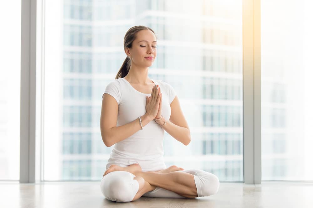 Woman wearing white does a yoga pose in front of a window.